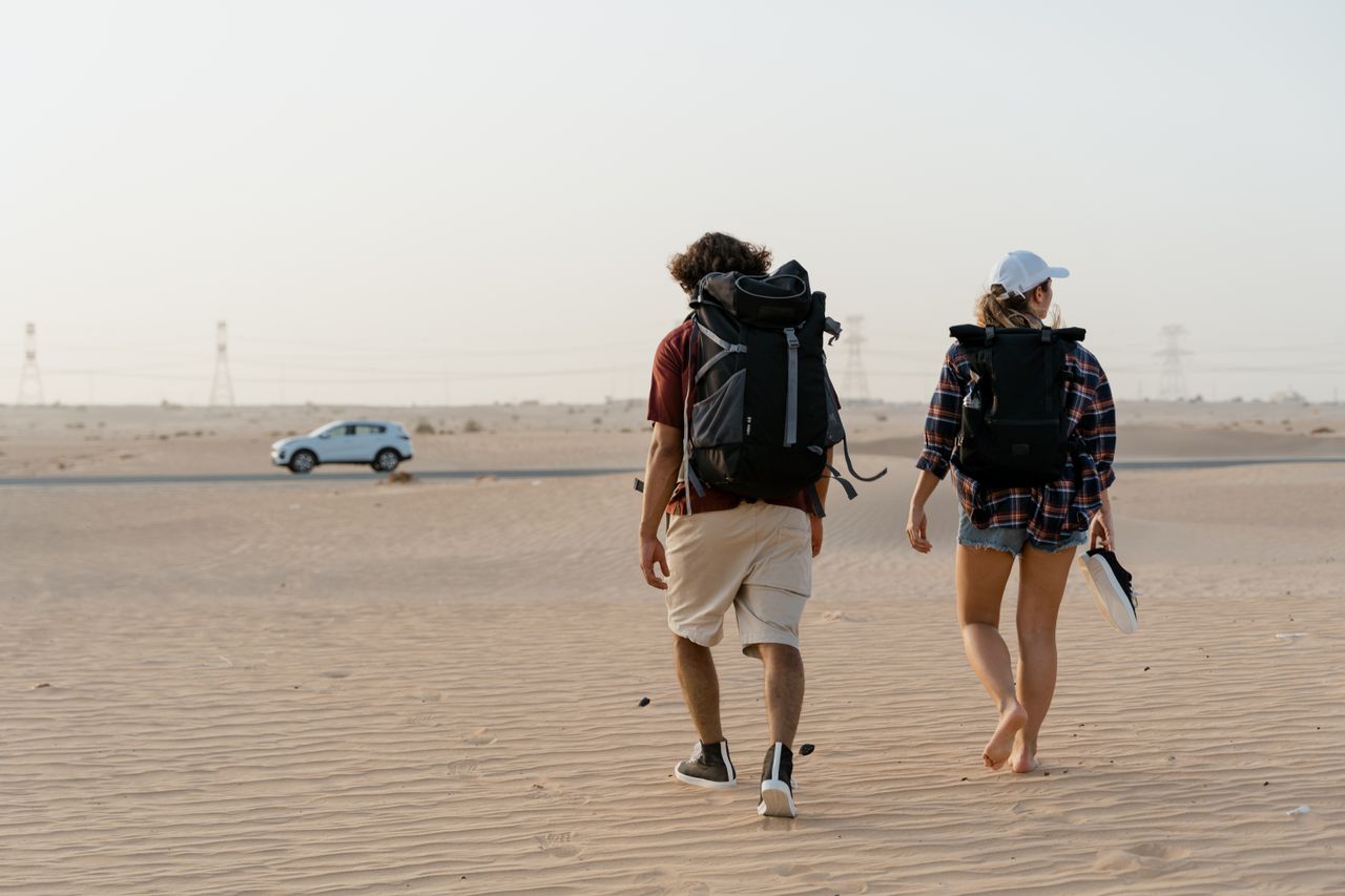 Contact A couple walking in a vast desert with backpacks, heading towards the horizon.
