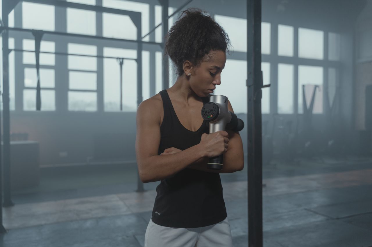 Contact Fit woman using a massage gun in a well-lit gym studio for muscle recovery.
