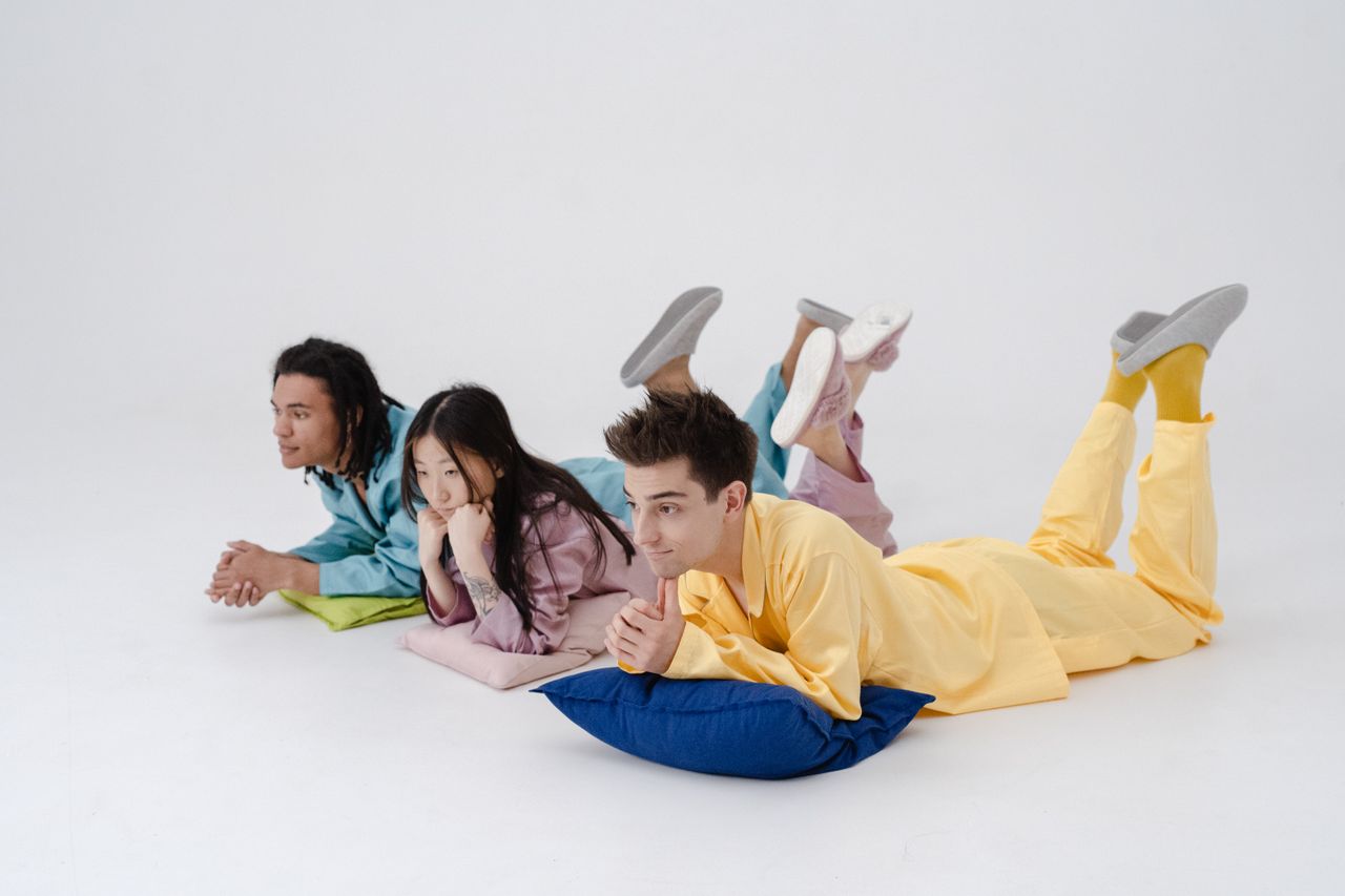 Contact Three young adults in colorful pajamas lying on pillows on a white surface, creating a cozy and relaxed atmosphere.